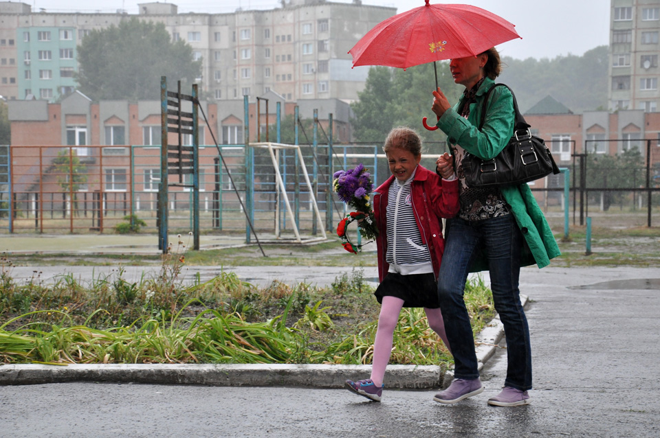 Донька із мамою поспішають на урочистості із традиційними квітами, прикриваючись парасолькою.