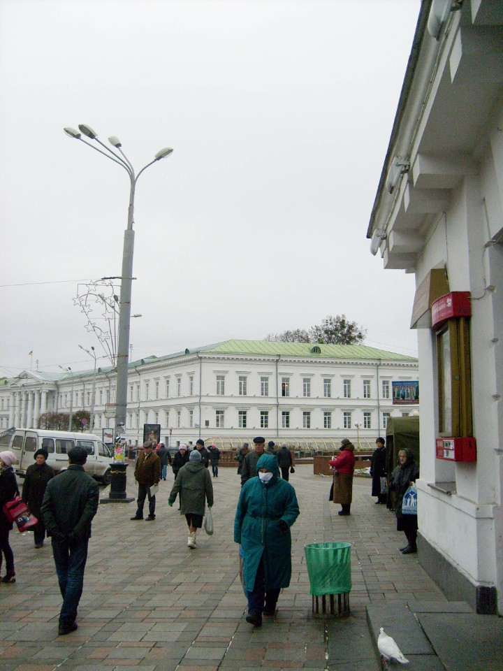 Зупинка громадського транспорту поблизу будівлі кінотеатру у листопаді 2009 року. Україна переживає епідемію грипу H1N1, а у Полтаві Олександр Мамай розпочинає передвиборчу кампанію (на фото жінка з пакетом ГО «Наш дім — Полтава»).